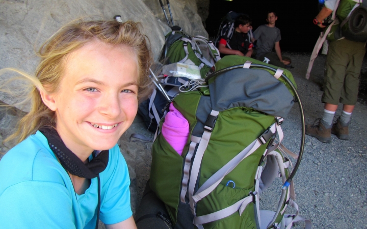 a young person sits beside their backpack and smiles at the camera